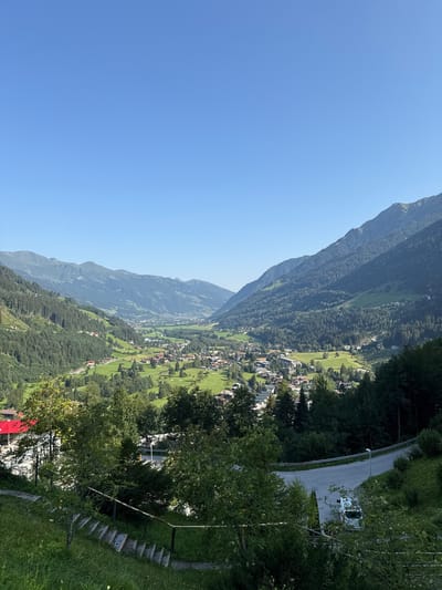 An aerial-view shot captures the picturesque Austrian town of Bad Gastein nestled within a verdant valley. The clear blue sky dominates the upper half of the frame, giving way to a panoramic vista of rolling green hills, densely forested slopes, and towering mountains in the distance. The town itself is a cluster of red-roofed buildings spread across the valley floor, surrounded by manicured golf courses and open green spaces. A winding road snakes through the scene, with a white van parked on its side. In the foreground, a set of concrete stairs descends amidst lush green grass and trees, suggesting a vantage point from a hillside overlooking the town.