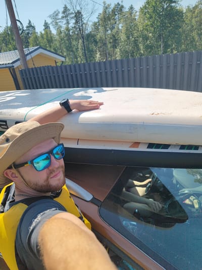 A man wearing a tan bucket hat, blue mirrored sunglasses, and a yellow life vest is taking a selfie while leaning on a white stand-up paddleboard. The paddleboard is placed on top of a vehicle. The man's left arm is resting on the paddleboard. The background shows a grey metal fence, a yellow building with a dark roof, trees, and a bright blue sky. The image is taken outdoors on a sunny day.