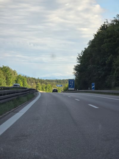 An asphalt highway, likely an Autobahn, is depicted in the vicinity of Berg, Germany. The road features at least two lanes in the direction of travel, marked by white dashed lines, with a solid white line defining the shoulder on the left. A metal guardrail runs along the left side of the road, behind which a white van is partially visible. Dense green forests flank both sides of the highway, extending into the background where faint blue silhouettes of mountains are discernible under an overcast sky with stratocumulus clouds.
In the distance, a dark-colored car is present in the left lane, traveling away from the viewer. Road signs are visible on both sides of the highway. On the left, a blue directional sign, partially obscured by trees, displays the letters "oge," consistent with a destination like Berg. On the right, a larger blue sign indicates service amenities with white pictograms for a gas station, food, and lodging. Further down on the right, a smaller blue sign with "100 m" likely indicates a distance marker. The scene is illuminated by ambient daylight, suggesting either an overcast day or late afternoon.