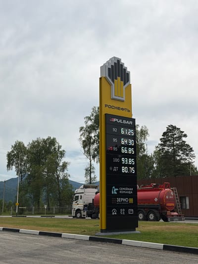 The image shows a "Rosneft" gas station in Ust'-Sema, Russia. A yellow sign displays fuel prices, with 92 octane at 61.25 rubles, 95 octane at 64.30 rubles, 95+ at 66.85 rubles, 100 octane at 93.85 rubles, and diesel (ДТ) at 80.85 rubles. Below the fuel prices, there are logos for "Pulsar," "Семейная команда" (Family Team), "Зерно" (Grain), and icons indicating 24-hour service and payment options. A white semi-truck with a red fuel tanker is parked to the right of the sign. The truck has "ОГНЕОПАСНО" (Flammable) markings. The environment is outdoor, with green grass, trees, and a cloudy sky. A road with black and white curbs is visible in the foreground.
