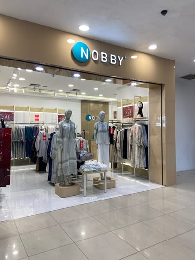 The image shows the exterior of a "NOBBY" clothing store in Kudus, Indonesia. The store has a beige facade with the "NOBBY" logo, featuring a blue circle with white lettering, prominently displayed above the entrance. The store entrance is a glass-fronted display showcasing mannequins dressed in modest fashion, predominantly long dresses and headscarves, in muted colors. Racks of clothing are visible inside, with several red "SALE" signs indicating promotions. The interior lighting is bright, illuminating the merchandise. The floor inside the store is tiled in white, while the surrounding area outside appears to be tiled in a light beige stone. The store is situated in what appears to be a shopping mall or commercial complex, given the indoor setting and the presence of ceiling lights and what looks like a security camera.