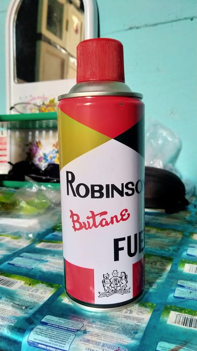 An upright, full-color, medium-shot photograph captures a can of "ROBINSON Butane Fuel" positioned on a blue, patterned surface that appears to be a table or counter. The can features a red cap and a white label with bold black lettering for "ROBINSON" and red script for "Butane," followed by "FUEL" in large black letters. A small crest with the words "ROBINSON ROBINSON" is visible near the bottom of the label. The background is out of focus, showing a mint-green wall and a partial reflection of a mirror. To the left of the can, out-of-focus objects include a green shelf and a floral-patterned container. The surface beneath the can is covered with multiple sheets of blue packaging, some with visible text and barcode patterns, potentially indicating a product display or storage area. The lighting suggests an indoor environment, possibly a shop or home in Banyusri, Indonesia. No people are present in the image.