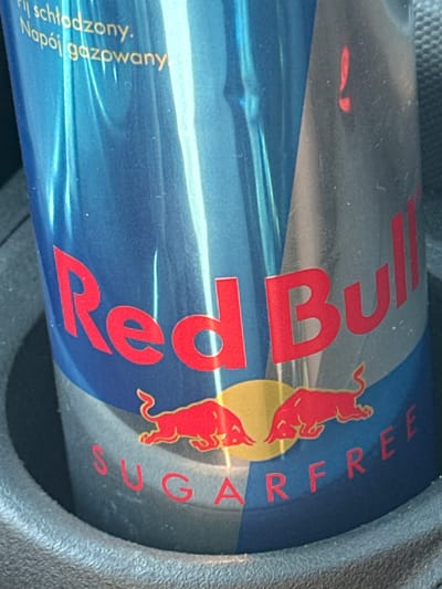 The media file shows a close-up, angled shot of a Red Bull Sugarfree energy drink can. The can is primarily blue with the iconic red Red Bull logo. The word "RedBull" is prominently displayed in large red letters, with two red bulls depicted on the can, one above the other, superimposed on a yellow circular background. The text "SUGARFREE" is also visible in red lettering.

On the lower left side of the can, there is text in a smaller font, written vertically. This text appears to be in Polish and reads "Napój gazowany. Najschłodzony." which translates to "Carbonated drink. Chilled."

The can is situated in a dark, textured holder, likely a cup holder in a vehicle, suggesting it is being transported or stored. The lighting is artificial, creating reflections on the metallic surface of the can. No people are visible in the frame. The scene is focused entirely on the product, with the background blurred and out of focus. The overall impression is that of a product shot or a casual snapshot of the energy drink. Given the location context of Rutesheim, Germany, this is likely a consumer in Germany.