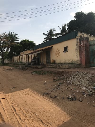 The image shows a street scene in Oko Erin, Nigeria. A weathered, single-story building with a pale yellow facade stands adjacent to a dirt road. The building has a green metal roof, a small square window, and decorative geometric patterns on the wall. The address "2 YOLA RD." is visible on the building's front. A small covered porch area is supported by posts.
The road is sandy with visible tire tracks. Debris and loose rocks are scattered along the roadside. Trees, including palm trees, are visible along the street, with power lines running overhead. The sky is overcast. There are no visible people or signs of recent human activity.