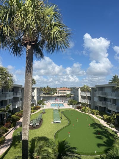 The image shows an aerial view of a resort or condominium complex in Hillsboro Beach, Florida, USA. The buildings are multi-story with balconies. There is a putting green with small red flags on it, a shuffleboard court, and a swimming pool area with lounge chairs. Lush greenery and palm trees surround the area. The sky is blue with scattered white clouds.