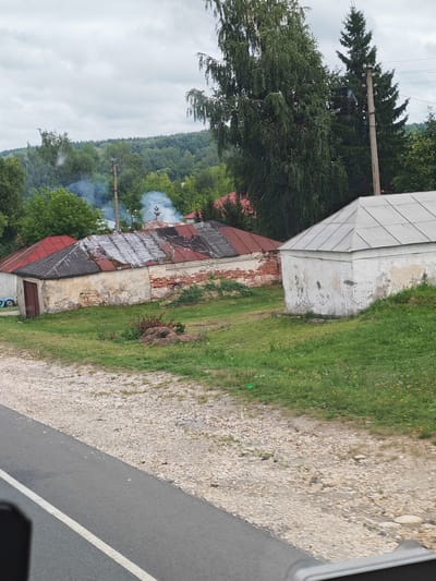 The image depicts a rural scene in Yambirno, Russia, featuring two old, dilapidated buildings with damaged roofs. One building, constructed from brick and plaster, shows significant decay and has a rust-colored, partially damaged metal roof. Smoke emanates from its chimney, suggesting a fire or heating activity. Adjacent to it is a smaller, white-washed structure with a grey, angular roof, also showing signs of age and disrepair. The surrounding area is overgrown with grass and dotted with small yellow flowers. A gravel path separates the buildings from a paved road, indicated by a white line. In the background, a dense forest stretches across rolling hills under a cloudy sky, with tall trees, including a birch and a fir, visible near the buildings and a utility pole with wires. The overall impression is one of rural neglect and the passage of time.
