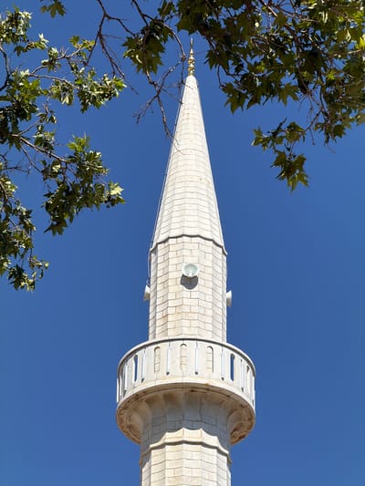 This low-angle outdoor, daytime shot shows the top portion of a minaret against a clear, deep blue sky. The minaret is constructed of light-colored stone blocks and has a conical roof topped with a gold finial. Around the middle of the visible portion of the minaret, there is a circular balcony with a white railing. Several white speakers are visible mounted on the sides of the minaret. In the upper left and top of the frame, green leaves and branches from a tree partially obscure the view, suggesting the minaret is surrounded by foliage. The scene is likely located in Ulcinj, Montenegro, a city known for its historical mosques and minarets.