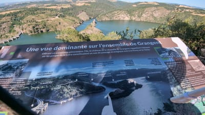 The image shows an informative sign with a landscape view of the Loire Gorges in Chambles, France. The sign has text in French and depicts a panoramic view of a river, hills, and a dam. The text describes the dominant view over the Grangent ensemble, mentioning the island, castle, and chapel. It also refers to the "Gorges de la Loire" and the historical significance of the area as a strategic passage. Labels on the sign point to various features in the landscape, such as "Vers Saint-Just," "Saint-Rambert," "Le Barrage" (the dam), "Signal de Grangent," "Lit de la Loire Courante" (riverbed), "Île de Grangent" (Grangent Island), "Maison de la Réserve Naturelle Régionale" (House of the Regional Nature Reserve), "Vers le Vieux Bourg et la Plage de Saint-Victor sur Loire" (Towards the Old Town and the Beach of Saint-Victor sur Loire), and "Les Camaldules." There are also small photographic inserts on the sign showing historical and modern views of the area, including the dam construction and surrounding landscape. The overall scene captured in the photograph is an outdoor viewpoint overlooking a vast body of water formed by the dam, with forested hills rising around it. The sign itself is the primary subject of the foreground.