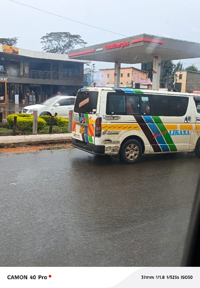 On a wet street, a white van with colorful stripes and decals is parked next to a sidewalk with greenery. The van's signage includes "LIKANA," "EXEDY," and "CHICKS.COM." In the background are a TotalEnergies gas station, a building with businesses, and a white car. The image is captured by a CAMON 40 Pro at 37mm, f/1.8, 1/525s, ISO50.