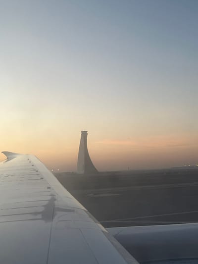 An airplane wing is visible in the lower right corner, with a view of a building resembling a control tower in the distance. The horizon is hazy, with a gradient of blue sky transitioning to orange, likely indicating dawn or dusk. The ground below appears to have an airport runway.