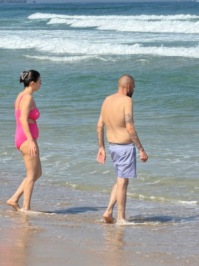 In Ashkelon, Israel, two individuals are depicted on a beach with the sea in the background. A woman, seen from the side, wears a bright pink swimsuit and has her hair pulled back with a clip. She is walking near the water's edge. Beside her, a man, also seen from the side, is shirtless and wears blue and white striped swim shorts. He also appears to be walking along the shoreline. The ocean displays multiple rows of white-capped waves, suggesting moderate surf. The sky is a clear blue, and a distant pier or structure is visible on the horizon. The sand is wet where the waves are reaching the shore.