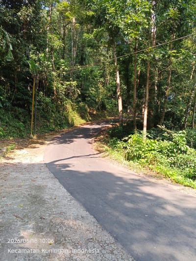 The image shows a narrow, paved road winding through a lush, green forest. The road is on the left side of the image, with a concrete sidewalk and some fallen leaves. The forest is dense with trees, foliage, and sunlight filtering through the canopy. A yellow pole with a black top stands to the left of the road. The timestamp "2026/06/18 10:06" and the location "Kecamatan Kuningan, Indonesia" are visible in the bottom left corner of the image. There are no people or animals present. The scene appears to be a quiet, natural setting in the Kuningan district of Indonesia.