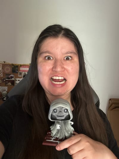 A woman with long dark hair holds a small figure of a hooded skeleton near her face. She is indoors with a corkboard covered with photos and cards visible in the background. The exact location within Câmara de Lobos, Portugal, cannot be determined from the image.