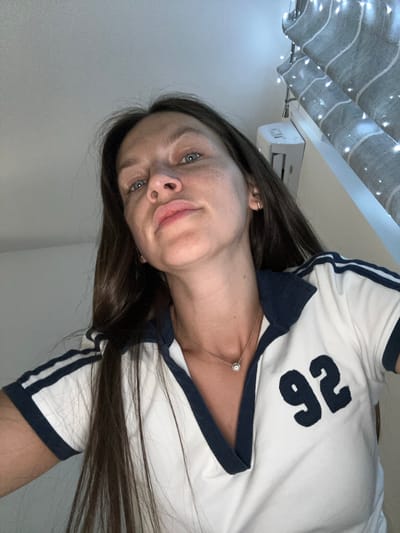 In a close-up, low-angle shot taken in a room, a woman with long, dark hair and blue eyes looks directly at the camera. She is wearing a white polo shirt with navy blue trim on the collar, sleeves, and a large "92" applique on the right side of her chest. A delicate silver necklace with a circular pendant is visible around her neck. The background shows a grey wall, a white air conditioning unit, and a window covered by striped blinds with string lights draped across them. The scene suggests an indoor setting, possibly a bedroom or living area, in Donji Pobori, Montenegro.