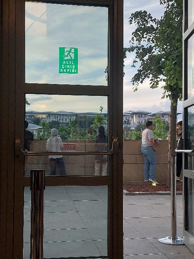 The image shows a view through a glass door, framed by brown metal. The door is open, revealing a view of the city. The door has a green emergency exit sign, reading "ACIL CIKIS KAPISI", on the top pane. The door's bottom pane shows a tiled floor. Outside the door, there are two people standing on a sidewalk, one man in a gray shirt and blue jeans, and one woman in a gray shirt and jeans. The man is facing the camera and the woman is looking to the right. The woman appears to be talking to another person, partially obscured by a pole. The man is facing forward. The sidewalk is gray and tiled. The area behind the sidewalk is composed of a tall concrete wall. In the distance, there are low-rise buildings. A tree with green leaves is visible on the right side of the image.  The location is Ankara, Türkiye, at latitude 39.925 and longitude 32.8.
