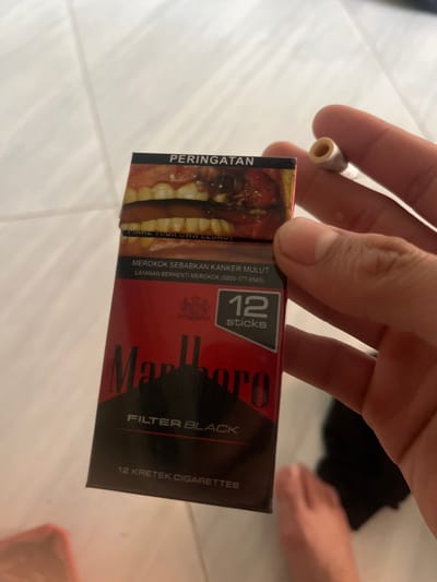 A person's hand is holding a cigarette and a pack of Marlboro Filter Black kretek cigarettes. The cigarette in hand is partially lit and appears to have been smoked. The pack is predominantly black and red, with the Marlboro logo clearly visible. Warning labels in Indonesian are present on the pack, including a graphic image of diseased teeth and gums, and text that translates to "WARNING: SMOKING CAUSES ORAL CANCER" and "QUIT SMOKING SERVICE (0800-177-6565)". The pack also indicates it contains "12 sticks" and is "12 KRETEK CIGARETTES". The scene appears to be indoors, with a tiled floor and what looks like a person's legs in the background. The lighting suggests it is daytime. The overall context points to a candid moment of someone holding cigarettes, with the prominent health warnings on the pack being a significant visual element.