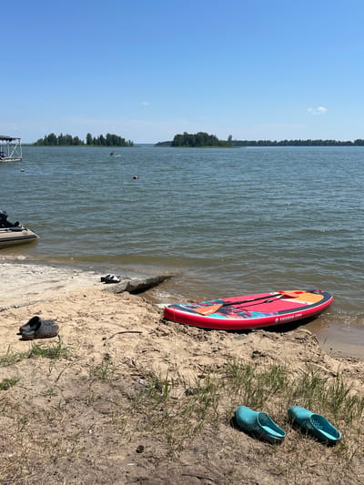 On a sunny day at the shore of a body of water, a red and blue stand-up paddleboard is partially submerged in the water next to a sandy beach. Several pairs of shoes are scattered on the sand: a black pair of Crocs is near the water's edge, and a pair of light blue perforated slip-on shoes are further up on the sand. In the background, a small inflatable boat is docked, and a person is seen paddleboarding on the water. The horizon is lined with trees, and the sky is clear blue. The location is the city of Borovoye, Russia.