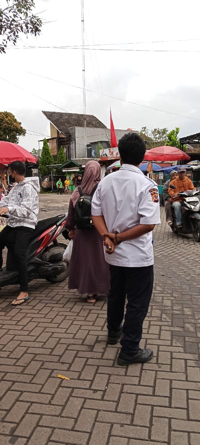 The image depicts a street scene in Banjaran, Indonesia (latitude: -7.046, longitude: 107.581). A man in a white shirt with a "POLDA" patch, black pants and black shoes is standing in the foreground. He has his hands clasped behind his back. In the background, a woman in a brown hijab and long brown skirt is standing. She is wearing a brown backpack and holding a white plastic bag. To the left of the image, another man, wearing a gray sweatshirt, black pants, and sandals, is standing next to a red and black motorcycle. He is holding a black helmet and a red and black object. In the background, there is a large metal tower, a multi-story building, and a street market with several stalls selling goods. There are also multiple people walking in the background. The street is paved with gray interlocking bricks, with some small green plants and a yellow leaf scattered on the ground. The sky is overcast and gray, with the sun obscured by clouds.