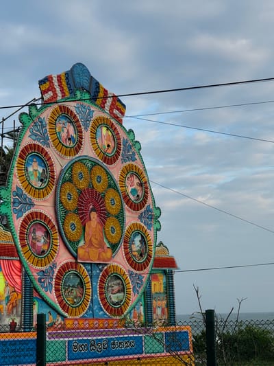 This image captures a close-up view of an ornately decorated structure, likely a festival float or temple decoration, against a cloudy sky. The central subject is a large, circular, multi-layered artistic piece adorned with numerous smaller circular panels, each containing painted scenes with figures. The dominant colors are pastel shades of pink, green, yellow, and blue, with intricate details and patterns. A prominent scene in the center depicts a figure in a saffron robe, possibly a religious icon. Other circular panels show various human figures in stylized settings.
The structure itself is elaborately designed with a scalloped edge and appears to be constructed from wood or a similar material, painted with vibrant imagery. To the right of the central piece, a vertical section of the decoration is visible, featuring rows of painted figures and what appears to be lettering in a non-Latin script. A green wire fence runs along the right edge of the frame, partially obscuring the structure and revealing a hint of greenery and a distant body of water or hazy landscape in the background. Electrical wires crisscross the sky, suggesting an outdoor setting. The lighting suggests it is daytime, with diffused light from the overcast sky. The overall impression is one of a colorful and culturally significant celebration or display. The visible text on the vertical panel is in Sinhala script.