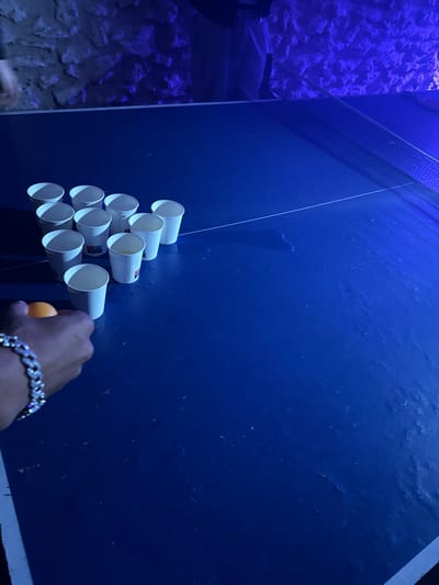 Scene in Palaiseau, France, features a blue backdrop where white cups are arranged in a triangular pattern. A person's hand, adorned with a silver bracelet, holds an orange ping pong ball near the cups. The environment includes a stone wall on the left side of the image. The lighting gives a blue cast to the scene.