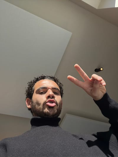 An individual is photographed in an indoor environment in Marche-lez-Écaussinnes, Belgium. The subject, a male with dark, curly hair and a beard, wears a black turtleneck sweater. He faces the camera directly, making a pouting expression with his lips. His right hand, positioned to the upper right of his head, displays a peace sign (V-sign) with the index and middle fingers extended. The background features a light-colored ceiling with angular planes and a single dark recessed light fixture. Illumination originates from an overhead light source, creating distinct shadows.