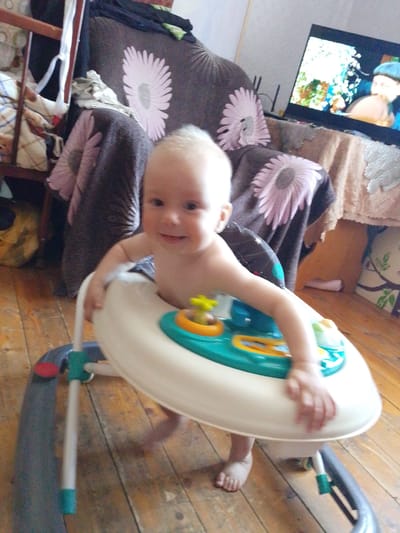 The image captures a close-up, low-angle view of a baby in a walker. The baby, with light-colored hair and a broad smile, is the central subject, looking directly at the camera with a joyful expression. The baby is wearing a walker, which is predominantly white with teal and yellow accents, and appears to be actively engaged with the toy features on the walker's tray. The baby's hands are visible, one resting on the walker's edge.
The setting appears to be an indoor environment, likely a living room, with wooden flooring. In the background, partially visible, is a sofa with a distinctive floral pattern featuring large pink flowers. To the right, a television screen is on, displaying an image with colorful visuals, suggesting entertainment is present. A portion of a wooden shelf unit with various items, including blankets and what seems to be a laundry basket, is visible on the left. The lighting suggests it could be daytime, but the indoor setting makes it difficult to ascertain specific weather conditions. The overall atmosphere conveyed is one of domesticity and a happy moment captured with a young child. No visible text is present in the image. The context provided places the location in Birsk, Russia.