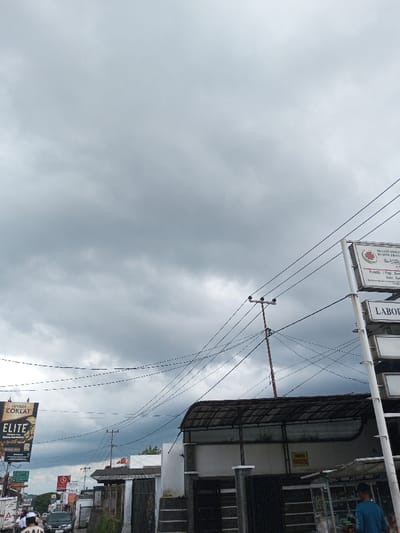 The image captures a street view under a heavily overcast sky in Cibadak, Indonesia. On the right side, a tall white pole supports multiple stacked rectangular signs. The top sign features a red and green logo, identifying "PRAKTEK DOKTER" (Doctor's Practice) and "KLINIK PRATAMA" (Primary Clinic). Below it, another sign reads "LABORATORIUM." Partially visible beneath these signs is a small vendor stall with merchandise behind glass, near which a male individual in a blue shirt and dark cap stands, facing away from the viewer. Behind these structures is a two-story building with a distinctive corrugated roof and a yellow sign bearing the word "GRATIS" (Free) on its white wall, fronted by a dark, segmented fence.

The central and left portions of the scene are dominated by a dense network of overhead electrical and communication lines crisscrossing the frame, connected to a prominent wooden utility pole. To the far left, a large vertical billboard displays an advertisement for "D'JAVA COKLAT ELITE," featuring a chocolate product. Below this, a bustling two-lane road is visible, populated by multiple automobiles and motorcycles. Several pedestrians are present, including a person in a white top visible on the far left. The street is lined with various commercial buildings, and faint outlines of distant hills or mountains are discernible beneath the cloudy horizon.
