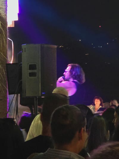 The image depicts an outdoor nighttime event with a stage in the background. A man with long, dark, curly hair and glasses is singing into a microphone. He is wearing a dark tank top. Behind him, a woman with dark, shoulder-length hair is visible, wearing a patterned top. In the foreground, several people's heads and shoulders are visible, suggesting an audience. The environment appears to be illuminated by stage lighting, with purple and white lights prominently featured. A large speaker is positioned to the left of the singer. The overall setting suggests a concert or performance in a public space in Balchik, Bulgaria.