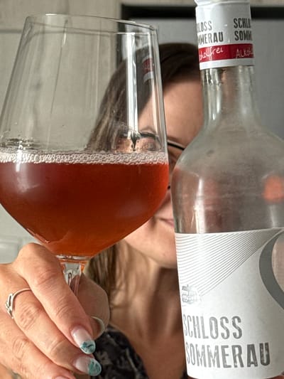 A person, partially obscured by a glass of reddish-brown liquid and a bottle, is in the foreground. The person is holding the glass with their left hand, which is adorned with a ring. The glass contains a frothy liquid. Beside the glass is a bottle with a white label featuring the text "SCHLOSS SOMMERAU ALKOHOLFREI" in black and red lettering. The background is blurred, suggesting an indoor setting. There are no discernible details about the location in Mesogi, Cyprus.