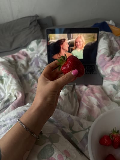 The image depicts a cozy, high-angle view of a person relaxing in bed in what appears to be Fehraltorf, Switzerland. The primary focus is a hand, featuring pink nail polish and a silver chain bracelet on the wrist, holding a large, vibrant red strawberry. The strawberry is held by its green leafy stem. To the upper right, a white bowl containing several other red strawberries is partially visible.
In the mid-ground, a silver laptop rests on the bed, its screen displaying a video or image featuring two individuals. One person on the screen has blonde hair and a discernible smile, while the other has dark, curly hair and is looking upwards. The laptop is positioned on patterned bedding, which features a floral design with pink, purple, and green elements against a light background. A plainer grey sheet or duvet is visible towards the bottom left. The overall scene conveys a relaxed and leisurely atmosphere, suggesting the person is enjoying fresh fruit while watching content on their laptop. No discernible text is visible within the image.