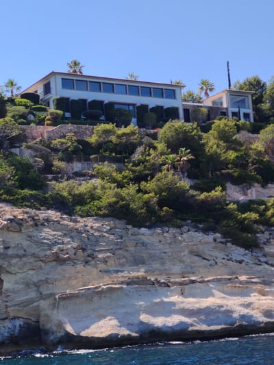 The image depicts a luxury villa complex situated on a steep, rocky hillside overlooking the Mediterranean Sea. The architecture is modern, with clean lines and large windows, suggesting a high-end property. Lush green vegetation, including trees and manicured shrubs, covers the hillside, interspersed with stone pathways and retaining walls. The sea below is a vibrant blue, with small waves gently lapping against the rocky shore. The sky is clear and bright, indicating a sunny day. The villa appears to be located in a well-to-do coastal area, possibly in a resort town. Given the context provided, this villa is likely located in Calvià, Spain, a municipality known for its affluent coastal resorts and beautiful scenery. No people are visible in the image.