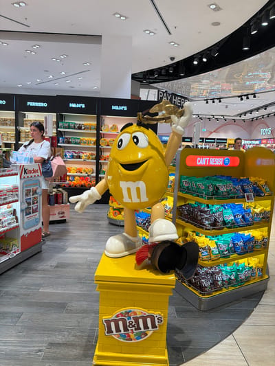 A life-size yellow M&M's character statue is positioned in the center of the frame, with its right arm raised as if holding an object. The statue stands on a yellow pedestal adorned with the M&M's logo. To the right of the statue, shelves are stocked with various M&M's candy bags. A woman stands to the left of the statue, looking towards the shelves. In the background, more shelves are visible, displaying other confectionery products, including Kinder and Ferrero brands. The scene appears to be an airport duty-free shop in Fiumicino, Italy, indicated by the "TOYS" sign and the general layout typical of such retail environments. The flooring is a dark wood-grain tile. Overhead, ceiling lights and a large digital screen displaying an advertisement are visible.