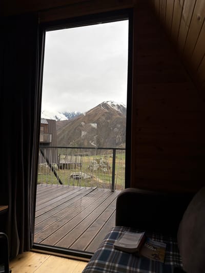 The photograph shows an interior view from a wooden cabin with a triangular roof, possibly in Sioni, Georgia, based on the book. The window shows a mountainous landscape with a cloudy sky, a wooden deck with a metal wire railing, and a building with windows. Inside the cabin, a portion of a couch with a patterned blanket and several books are visible. The lighting suggests daytime.