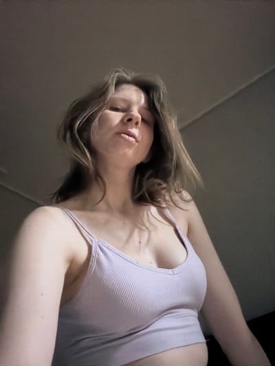The image shows a low-angle shot of a young woman from the chest up. She has light brown hair that falls around her shoulders and is looking upwards with her eyes partially closed and lips slightly parted. She is wearing a light purple ribbed tank top. The background appears to be an indoor setting with a textured wall and ceiling, dimly lit. There is no indication of the city of Dongducheon-si, South Korea in the image.