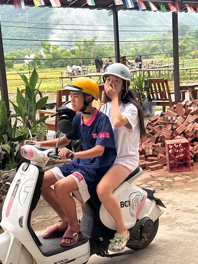 In Mai Chau, Vietnam, two young individuals are seated on a white electric scooter. The individual in front, wearing a yellow helmet and a blue polo shirt with white trim and a UNIQLO logo, operates the scooter. To the rear, a second individual with a grey helmet covers their mouth with their right hand while wearing a white t-shirt and light pink shorts. The scooter is parked on a dirt path, with a pile of red bricks to the right and lush green foliage and rice paddies in the background, indicative of a rural setting. A string of international flags hangs above the scene.