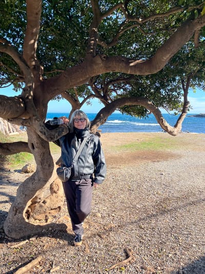 The image captures an adult female standing outdoors in Antibes, France, on a clear, sunny day. The individual is positioned slightly to the left of the frame's center, leaning her right arm against the gnarled, multi-trunked base of a large tree with dense green foliage. She has shoulder-length blonde/grey hair and wears rectangular glasses. Her attire consists of a black, long-sleeved jacket (appearing to be leather or faux leather), dark trousers, and dark closed-toe shoes with thick, light-colored soles. A black cross-body bag is slung over her left shoulder, resting at her left hip. She holds a partially filled clear plastic water bottle in her left hand. The ground beneath her is a mix of light brown gravel and small stones, with visible tree roots and sparse patches of green grass. In the background, an expansive blue body of water, likely the Mediterranean Sea, exhibits white-capped waves. A clear, light blue sky is visible above the horizon. To the far right, a dark rocky outcropping or peninsula extends into the sea. The scene is brightly illuminated by natural sunlight, casting defined shadows.