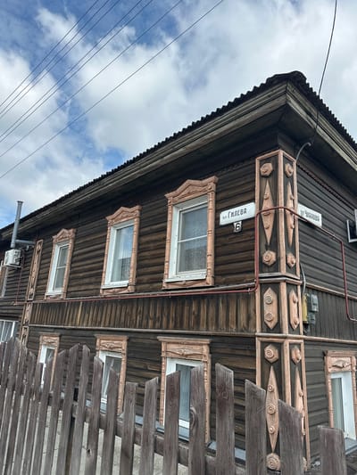 The image shows a close-up view of the corner of a two-story wooden house in the city of Nogornyi, Russia. The house is clad in dark horizontal wooden planks and features ornate pink-colored wooden trim around the windows and on the corner. Two street signs are visible on the corner post: one reads "пер. ГИЛЕВА 6" (Pereulok GILEVA 6, meaning GILEVA Lane, house number 6) and the other reads "ул. ЧКАЛОВА" (Ulitsa CHKALOVA, meaning CHKALOVA Street). A rustic wooden fence runs along the bottom of the image. The sky above is cloudy with visible power lines crossing the frame. An air conditioning unit and a ventilation pipe are mounted on the side of the house. The overall scene depicts a residential building in a Russian urban or suburban setting.