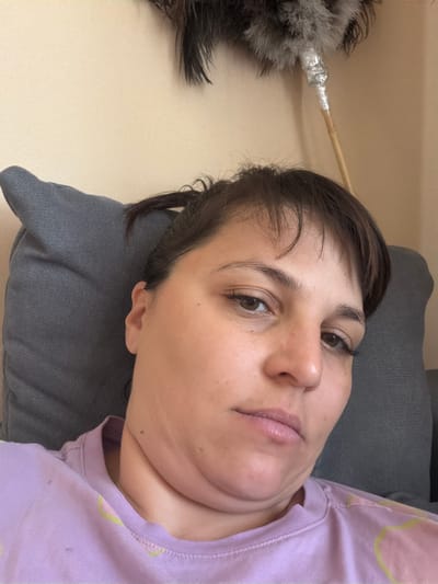 A woman with dark hair and brown eyes is lying on a grey pillow, with a faint smile on her face. She is wearing a light purple t-shirt. In the background, a feather duster with a metallic handle is visible against a plain wall. The scene appears to be indoors, possibly in a home in the city of Vladaya, Bulgaria.