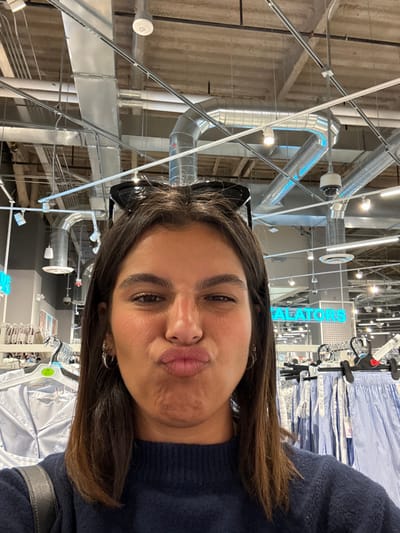 A young adult female is depicted in a close-up, chest-up selfie, positioned centrally within a large retail clothing store. She has dark brown, medium-length hair with black sunglasses resting on top of her head, and wears a dark blue knitted sweater with a dark shoulder bag strap visible on her left side. She has multiple small hoop earrings in both ears. Her face displays a playful expression, characterized by pursed lips and squinted eyes.
In the background, light-colored collared shirts with subtle patterns are visible on white hangers to the left, and light blue and white striped bottoms are visible on black hangers to the right. An industrial-style exposed concrete ceiling features extensive silver-colored ventilation ducts, numerous overhead lights, and at least one white dome security camera. A turquoise illuminated sign is partially visible in the distance, displaying "-ALATORS," suggesting "ESCALATORS." This scene is located inside a Primark store, specifically at 1618 Chestnut Street in Philadelphia, Pennsylvania, United States.