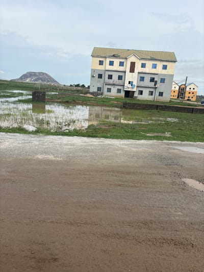 This image depicts an outdoor scene in Angwan Sayawa, Nigeria, featuring a multi-story residential building alongside a damp landscape under an overcast sky.

In the foreground, a rough dirt or gravel road occupies the right side of the frame, showing wet patches and small puddles. Adjacent to the road, a wide expanse of green grass is visible, heavily interspersed with large areas of standing water, suggesting recent rainfall or poor drainage.

The central subject on the left side of the frame is a prominent multi-story building, likely an apartment complex, with a facade that combines sections of light beige and grey. It features multiple rectangular windows with dark frames arranged on different floors. Several white satellite dishes are attached to the exterior of the building at various levels. The roof of the building appears to be a light, possibly greenish, color.

In the background, other residential structures, some with yellow or orange exteriors, are partially visible. A distinctive, rocky hill or small mountain rises in the far distance under a grey, cloudy sky. Utility poles or similar structures are also present in the background. The scene suggests a developing area with a mix of established buildings and open, somewhat undeveloped land. The time of day is daytime, with subdued lighting typical of overcast weather. No discernible text is visible in the image.