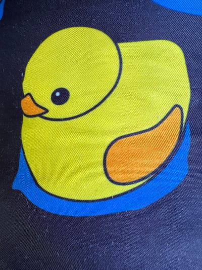 The media file is a close-up shot of a fabric with a printed design of a yellow cartoon duck. The duck is depicted in a simplified, playful style with a round body, a small head with a black eye and a bright orange beak. It is outlined in black. Part of its body is overlapped by a bright blue shape, which in turn is set against a dark navy blue background fabric. The texture of the fabric is visible, showing a woven pattern. The image appears to be taken in daylight, with natural lighting illuminating the colors and textures. There is no text visible in the image. The context provided places this image in Sant Feliu de Guíxols, Spain, though this specific location is not visually indicated in the photograph itself.