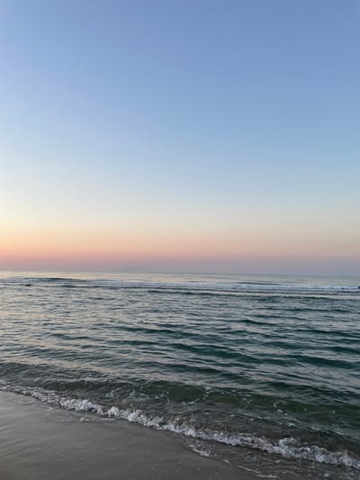 The image shows a tranquil beach scene at dawn or dusk. The sky transitions from a pale blue at the top to soft pink and peach hues closer to the horizon. A faint outline of a distant landmass is visible on the horizon. The sea is calm, with gentle waves lapping at the sandy shore. The water is a deep turquoise color, reflecting the colors of the sky. Foamy white water from the receding waves covers the sand at the edge of the sea. The sand itself appears wet and dark where the water has touched it, transitioning to a lighter, dry color further up the beach. No people or man-made objects are visible in the frame. The overall atmosphere is serene and peaceful. The location is described as a beach in an unknown city in Greece.