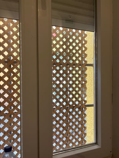 The image displays an interior perspective looking outwards through a white-framed window, likely a sliding type, with a partially retracted white roller shutter at the top. Immediately beyond the window glass, a light brown wooden lattice screen with a diamond pattern is visible, serving as a privacy barrier or decorative element. Through the apertures of the lattice, portions of a vibrant yellow stucco wall are discernible on the right side, along with a horizontal metallic bar, possibly part of a security grille or railing. Further visual information beyond the lattice suggests an urban environment, with subtle hints of green (possibly vegetation), blue (sky or reflections), and grey (pavement or ground) through the gaps. In the bottom-left corner, a segment of a clear plastic bottle with a blue cap is present on an interior surface. No individuals or ongoing actions are depicted. The architectural style, particularly the yellow stucco and lattice, is consistent with common residential or building aesthetics found in Sevilla, Spain; however, a precise location within the city cannot be definitively identified from the image alone.