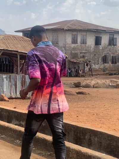The image depicts an outdoor scene in Egbedi, Nigeria, under an overcast sky. In the foreground, a male individual is walking away from the camera, positioned on a low concrete structure. He wears a short-sleeved collared shirt with a denim blue collar, featuring a gradient design from purple to pink to orange, adorned with dark palm tree silhouettes. He is also wearing dark, possibly distressed, jeans.
The ground throughout the scene is reddish-brown dirt, scattered with various rocks. To the left midground, a structure with a corrugated metal roof and walls is partially visible, with what appears to be laundry hanging inside. In the center midground, a makeshift shelter with a corrugated metal roof is present. Underneath this shelter, approximately five individuals, primarily men, are observed; some are seated, others standing, near a small white table or counter. A white standing fan is positioned outside the shelter.
In the background, a two-story concrete building with a visibly weathered and dirty facade and a rusted corrugated metal roof dominates the view. The building features multiple rectangular windows; some have closed wooden shutters, while others are open or appear empty. A distant hill or mountain is visible on the far left background.