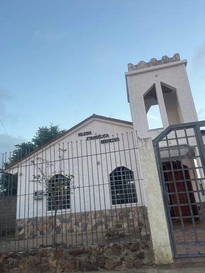 The image shows a white church building with a bell tower in the city of San Antonio, Venezuela. The church is identified by the Spanish words "IGLESIA EVANGÉLICA EBENEZER" above its entrance. A metal fence with a gate in front of the entrance partially obscures the building. The sky is clear and blue with some faint clouds. A small tree is planted in front of the church, between the fence and the building. The church is built with white stucco walls and a red tiled roof. The bell tower is square and has arched openings at the top. The gate is made of black metal bars and has a wooden door visible behind it. The base of the church wall is adorned with a mosaic of colorful stones. The overall scene depicts a religious building in a residential or semi-urban area, likely on a clear day.