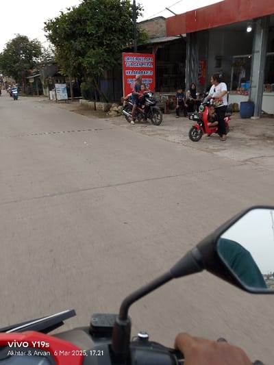 The image shows a street scene in Kecamatan Gabuswetan, Indonesia, taken from the perspective of a motorcycle rider. The rider is driving down the road towards a storefront on the right. The storefront has a large red sign that reads "GROSIR KELAPA TUA DAN MUDA, KELAPA PARUD GILIRU UMU" in Indonesian. There are two motorbikes parked in front of the store, and three people are standing near them. A red motorbike with a child on the back is moving towards the camera. The image is taken from the perspective of a motorcycle rider who is holding the handlebars. The road is paved and there is a black line painted in the center. The day is clear and the sun is shining.