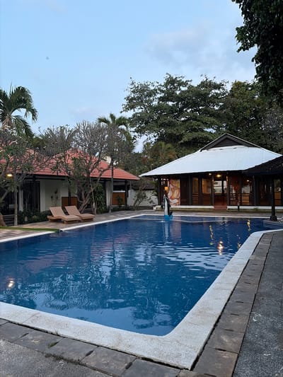 The image depicts an outdoor swimming pool area next to a building with a white, corrugated metal roof. The pool is filled with blue water, reflecting the ambient light and the surrounding environment. Two lounge chairs are placed on the paved area beside the pool, suggesting a place for relaxation. The building appears to be a resort or hotel, with multiple windows and open doorways revealing interior spaces. Trees with dense foliage and bare branches surround the area, contributing to a tropical or subtropical setting. Palm trees are also visible, further reinforcing the location's context. The sky is overcast with a light blue hue, indicating either early morning or late afternoon. A single person is visible near the edge of the pool, seemingly interacting with the water. The scene evokes a sense of tranquility and leisure, characteristic of a vacation destination. The location cues, such as the architecture and vegetation, along with the provided context of Banjar, Indonesia, confirm the setting.