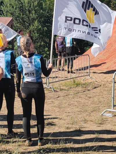 The image shows a group of people in athletic wear participating in an outdoor event, likely a race or competition. In the foreground, a woman with blonde hair in a braid is wearing a black long-sleeved shirt and black leggings, with a blue and white bib that reads "6517" and "ГОНКА ГЕРОЕВ". Another person, partially visible on the left, is wearing a similar blue and white vest over a black long-sleeved shirt and black leggings. A large white flag with black and yellow lettering is prominent in the background, displaying "РОСНЕФТЬ" and "ТОПЛИВНЫЕ". Other participants are visible in the background, some holding flags or wearing bibs with numbers like "1399" and "599". The environment appears to be sandy with sparse vegetation, suggesting an outdoor arena or a training ground. The presence of metal barriers indicates a demarcated course. Based on the context of the "Гонка Героев" (Race of Heroes) and "Роснефть" sponsorship, the event is likely taking place at the "Geroev Park" in Sertolovo, Leningrad Oblast, Russia, a known venue for such competitions. The lighting suggests it is daytime with sunlight casting shadows.