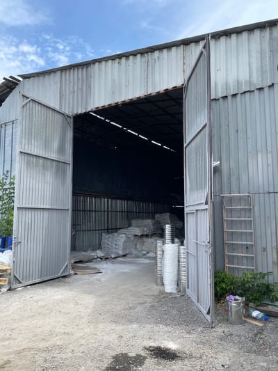 The image depicts the exterior of a corrugated metal warehouse with large, open double doors. The warehouse appears to be located in a semi-industrial or rural area, as indicated by the gravel ground and surrounding vegetation. Inside the open doorway, stacks of white bagged materials, possibly construction supplies or agricultural products, are visible. Several cylindrical objects, also white and wrapped in plastic, are stacked in front of the warehouse entrance. To the right of the entrance, a metal ladder is propped against the exterior wall, and a silver bucket with a purple object inside and a plastic bottle are on the ground. The sky above is partly cloudy. There are no people visible in the image. The overall scene suggests a storage or distribution point for goods.