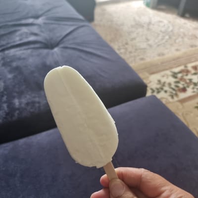 A close-up, medium shot shows a hand holding a white popsicle. The popsicle is elongated and slightly curved, with a visible indentation down its center. The hand appears to be that of an adult, with visible fingerprints. The popsicle is held at an angle in front of a dark blue upholstered surface, possibly a couch or chair, which takes up the lower left and upper left portions of the frame. Beyond the blue surface, a beige carpet with a subtle floral pattern is visible, extending across the right side of the image. The lighting suggests an indoor setting, possibly during daylight hours due to the slight brightness and shadows. The overall scene appears to be a casual, domestic environment. There is no discernible connection to the city of Aktobe, Kazakhstan.